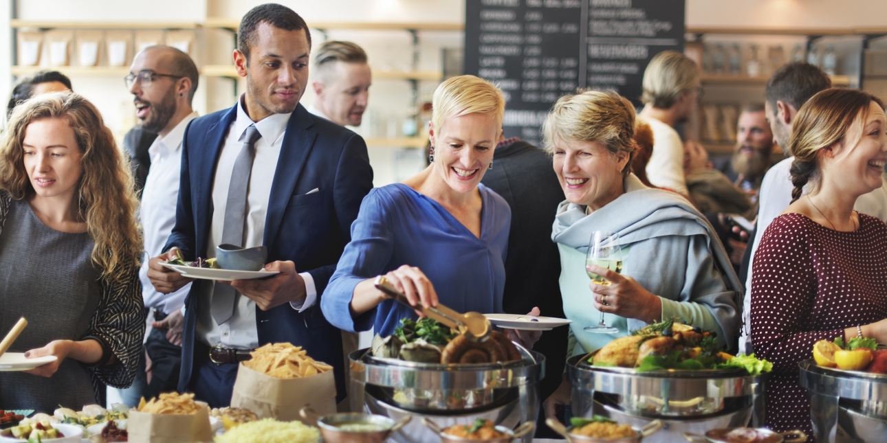 A dozen smartly dressed people mingling at an indoors event. In the foreground are four white woman and one mixed race man holding plates and glasses and reaching into a full buffet of assorted food